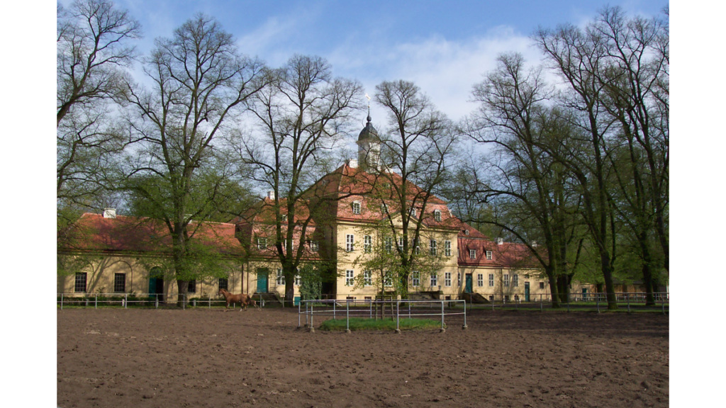 Ein dreistöckiges Gebäude hinter Bäumen und einer Pferdekoppel