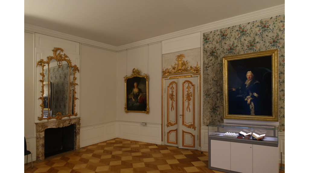 Ein edles Zimmer im Schloss Schönhausen mit einer mit gold verzierten Tür und zwei Portraits der Königin, außerdem ein ebenfalls mit Gold verzierter Spiegel