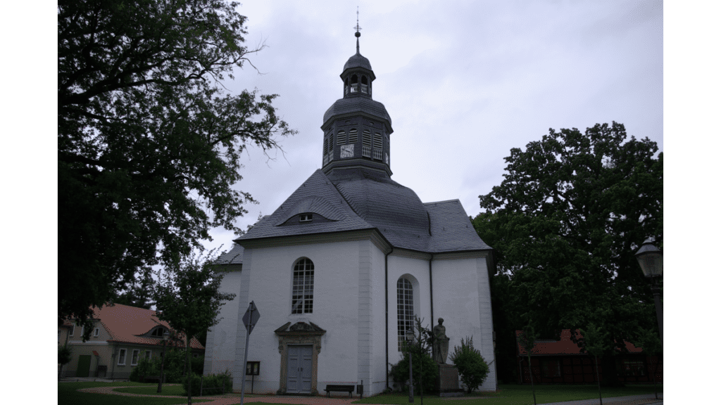 Der Blick auf eine weiß gehaltene Kirche zwischen Bäumen mit einem kleinen Glockenturm. Das Dach ist aus grauem Schiefer.