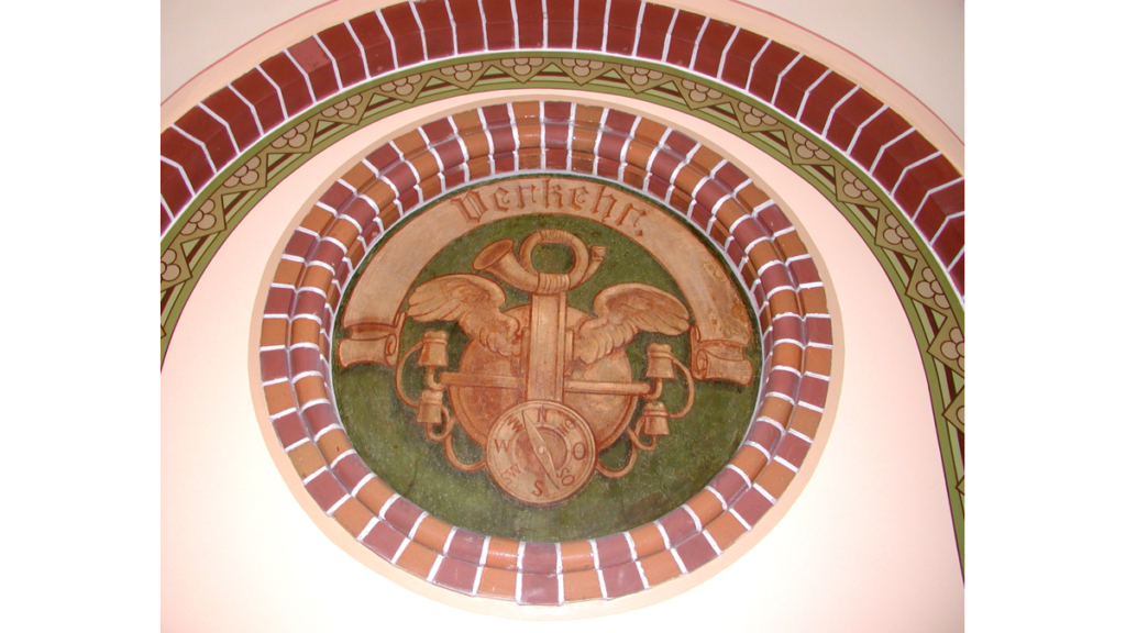 Ein in der Mauer eingelassenes, rundes Medaillon mit der Beschriftung "Verkehr" und einem Posthorn und einem Kompass