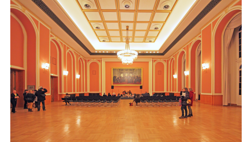 Ein großer in Terrakottatönen gehaltener Saal im Roten Rathaus, der Große Festsaal. Ein Kronleuchter hängt an der Decke und Stühle für ein Publikum sind aufgestellt; im Hintergrund spielt ein Kammerorchester.