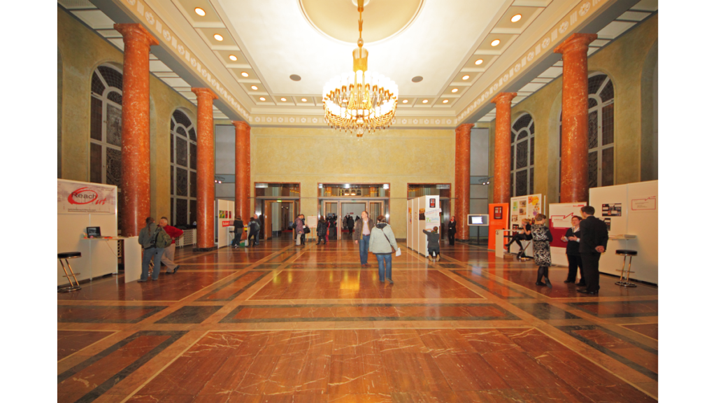 Ein großer von Marmorsäulen umrahmter Saal mit Parkettboden, an der Decke hängt ein Kronleuchter. Es handelt sich um den Wappensaal des Roten Rathauses.