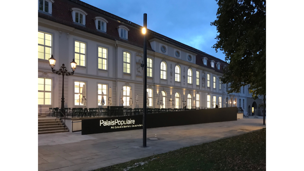 Ein im Dunkeln beleuchtetes, zweistöckiges, klassisches Gebäude, vor dem das Schild "Palais Populaire - Art, Culture and Sports by Deutsche Bank" steht.