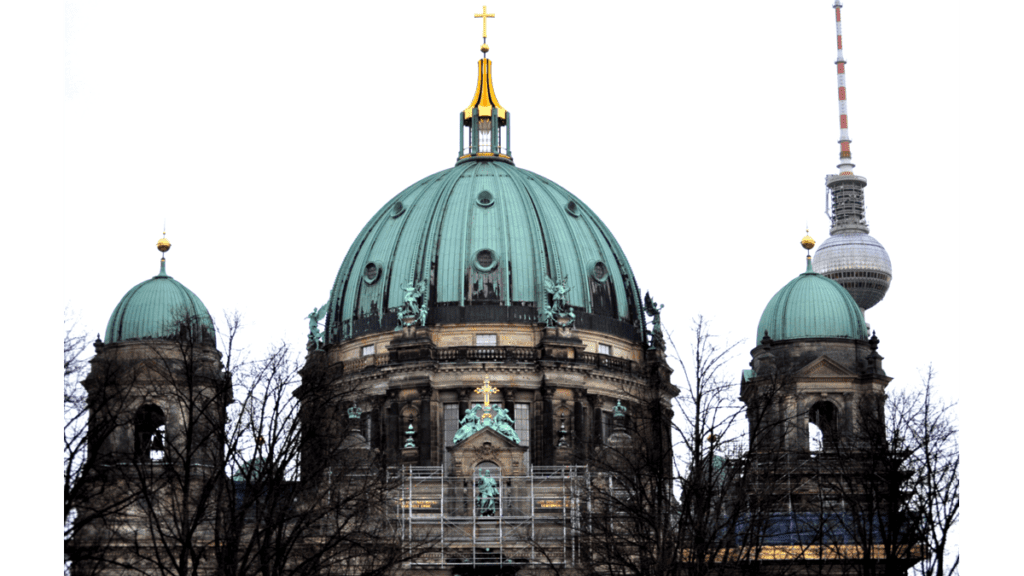 Die Vorderansicht des Berliner Doms mit der Hauptkuppel in der Mitte und zwei kleineren rechts und links davon. Im Hintergrund der Fernsehturm