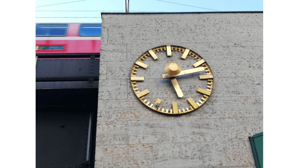 Eine Bahnhofsuhr am Bahnhof Zoo zeigt die Uhrzeit "12 Minuten nach Fünf" an. Im Hintergrund fährt ein Zug.