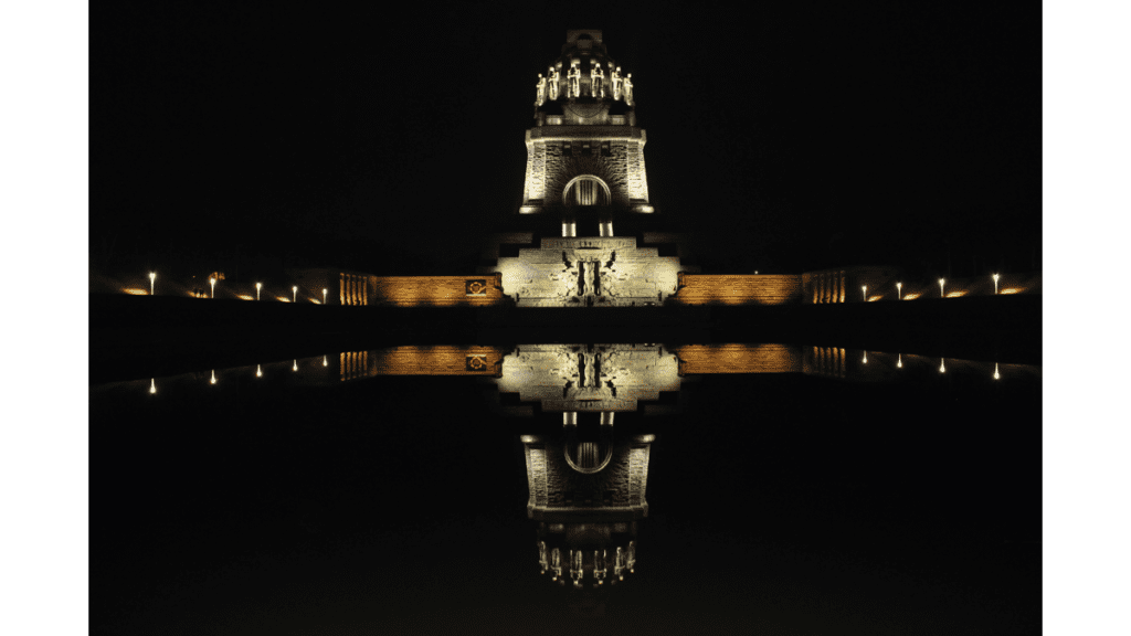 Das Völkerschlachtdenkmal bei Leipzig hell erleuchtet in der Nacht. Es wird im davorliegenden See gespiegelt.