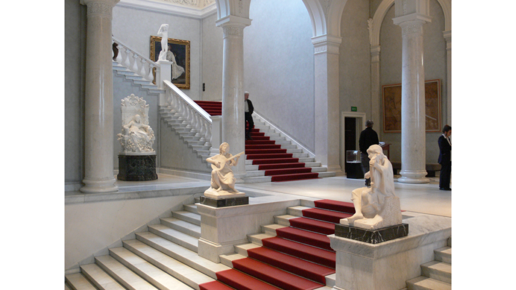 Das im klassischen Weiß gehaltene Treppenhaus der Alten Nationalgalerie mit einem dunkelroten Teppich und verteilten Statuen
