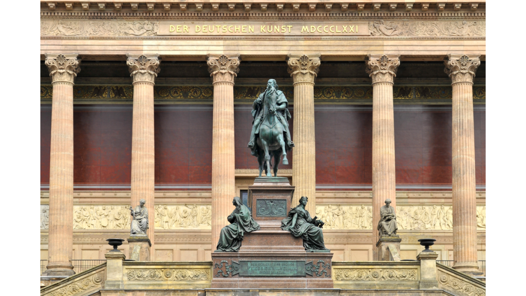 Das Reiterstandbild Friedrich Wilhelms IV. vor den Säulen der Fassade der Alten Nationalgalerie. Zu dessen Füßen sitzen zwei allegorische Frauenfiguren.