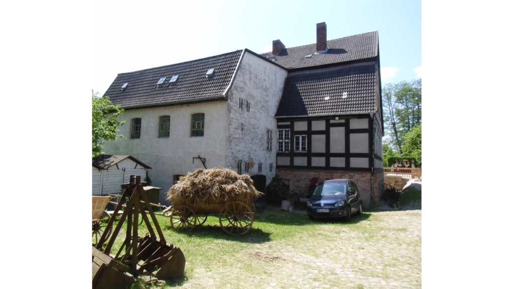 Die Klostermühle Boitzenburg von der dem Wasser abgeneigten Seite. Ein Heuwagen steht vor dem teils mit Fachwerk ausgestatteten Gebäude - neben einem Auto