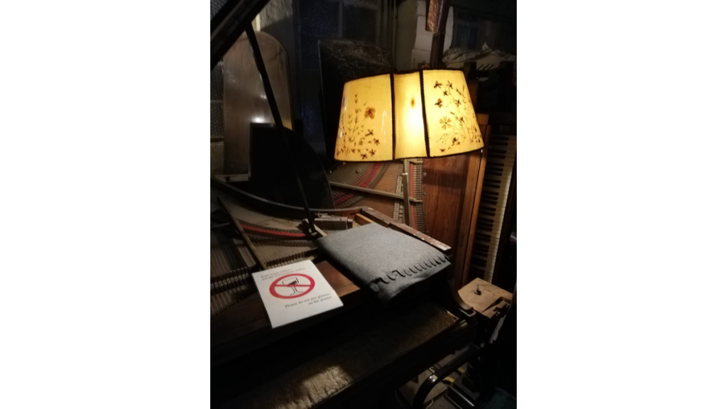 Eine Stehlampe neben einem Piano, auf dem eine Decke und eine Warntafel liegt, mit der gebeten wird,sein Glas bitte nicht hier abzustellen.