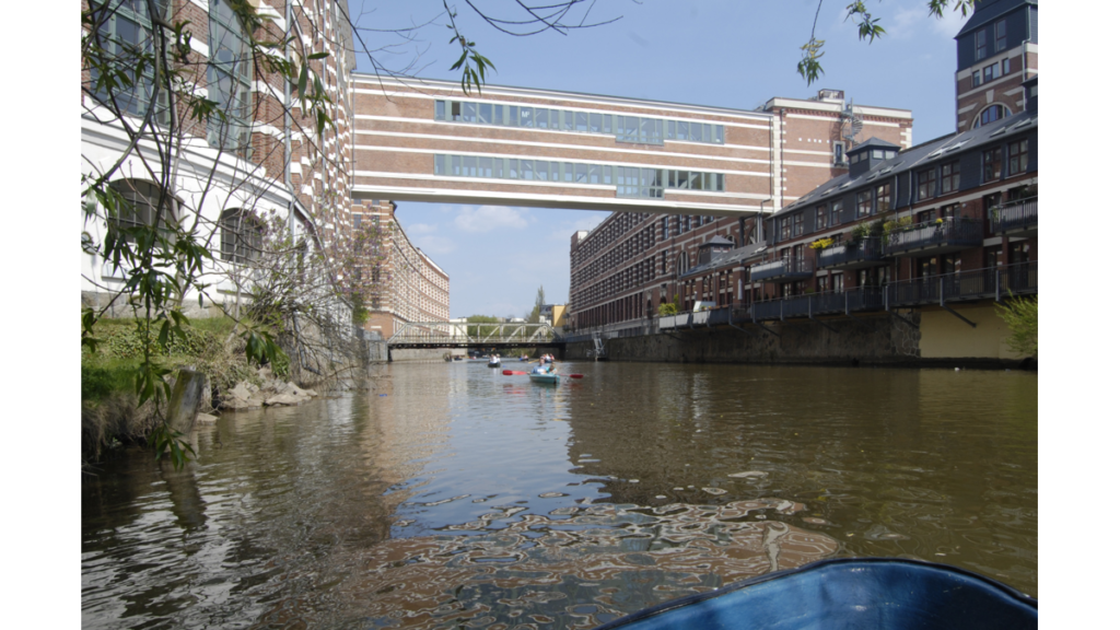 Die Buntgarnwerke vom Wasser aus. Eine Gebäudebrücke über zwei Etagen verbindet die Gebäude links und rechts vom Fluss.