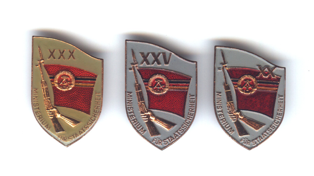 Drei Orden des Ministeriums für Staatssicherheit mit der DDR-Flagge und einem gehaltenen Gewehr