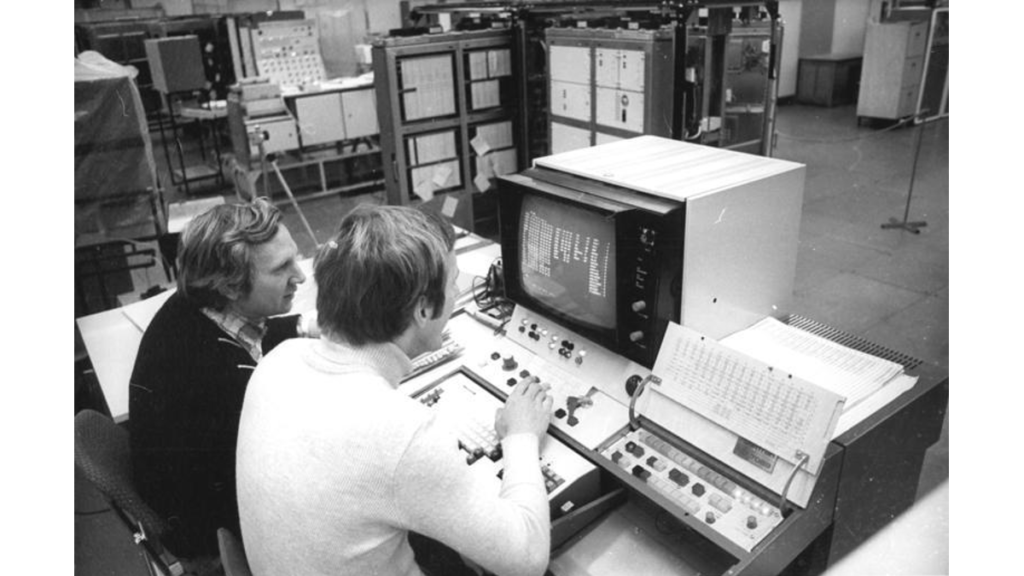 Ein schwarz-weiß Foto, in dem zwei Männer vor einem Computer sitzen und ein Programm studieren